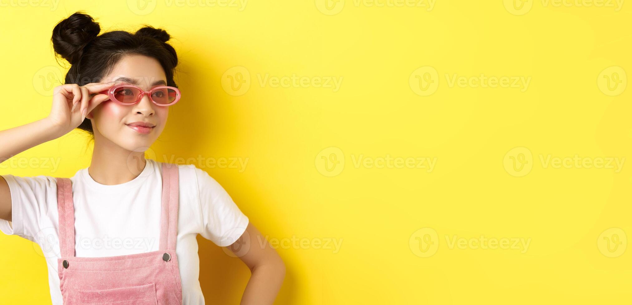 Beautiful asian girl put on stylish sunglasses and smiling, wearing trendy summer outfit, standing on yellow background photo
