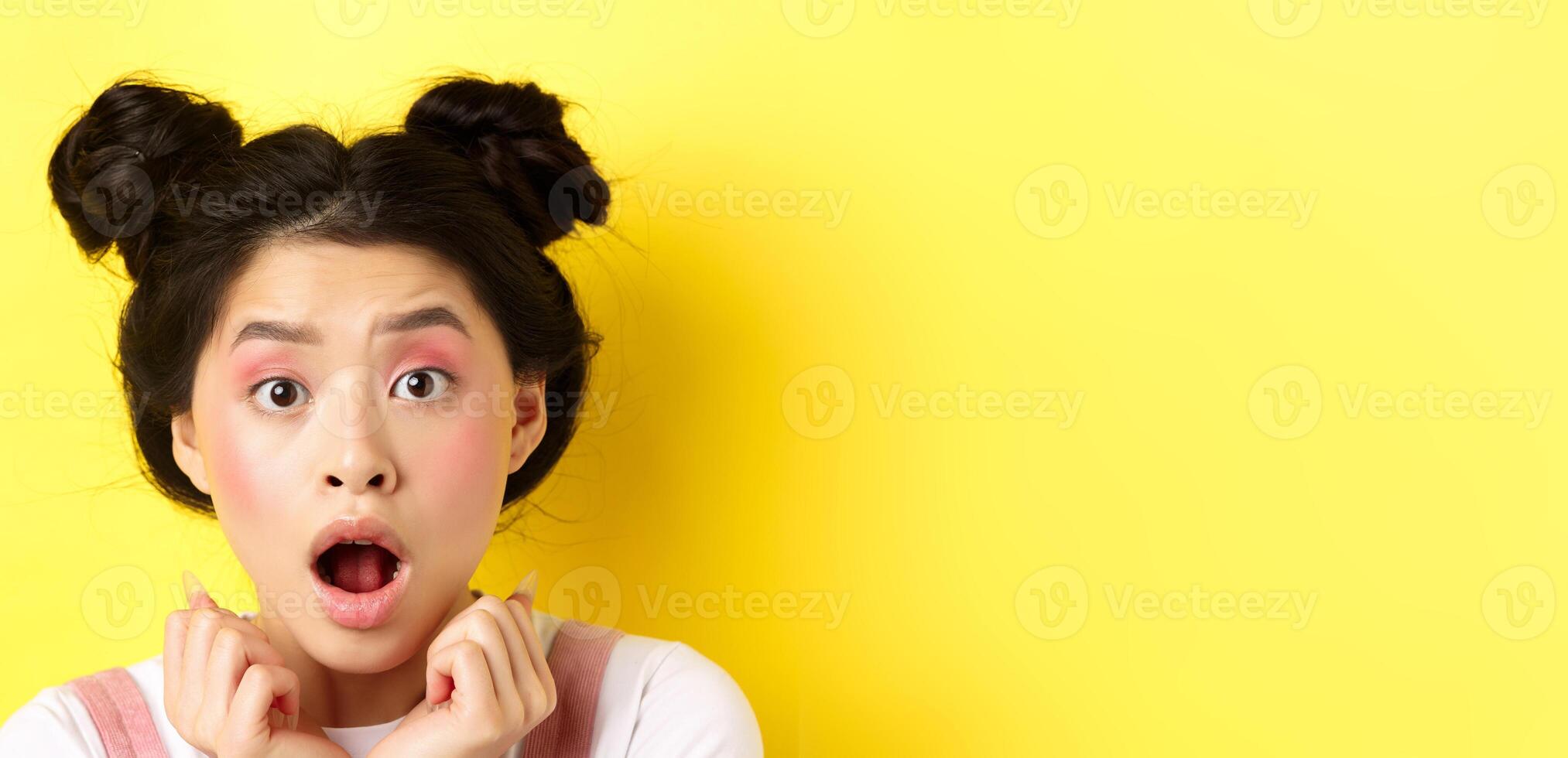 Head shot of shocked asian girl with hairbuns and glamour makeup, open mouth and looking startled at camera, standing against yellow background photo