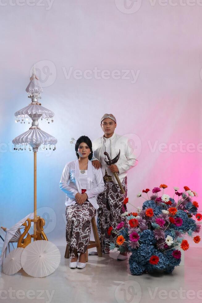 full body pair of lovers, a young Asian man and woman, sitting elegantly next to a man carrying a keris wearing modern Javanese traditional clothing, a white velvet bridal kebaya on a white background photo
