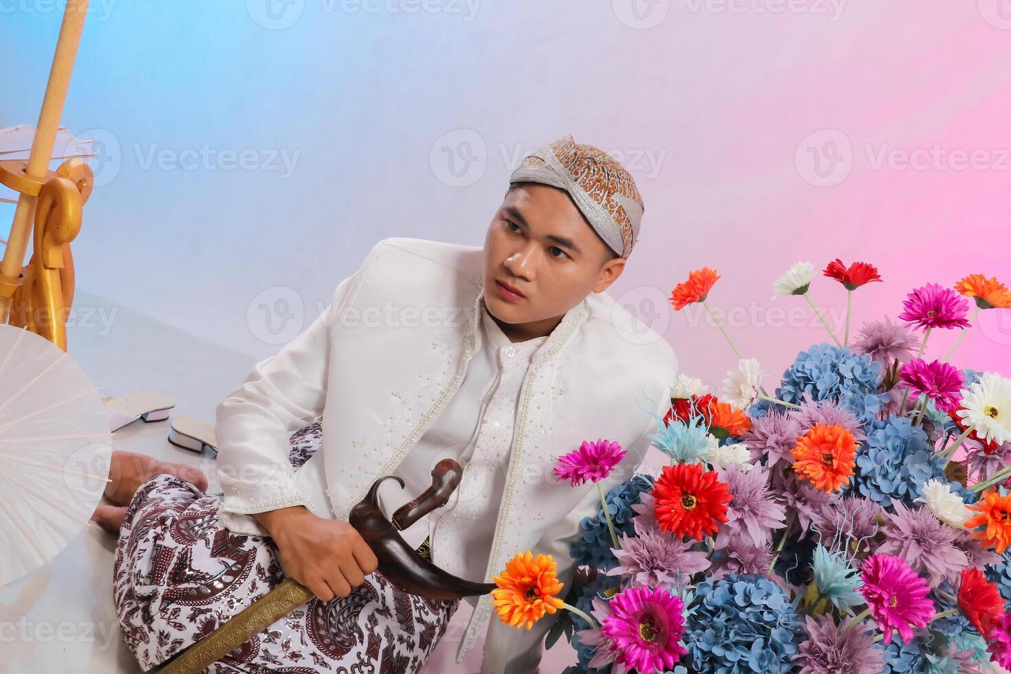 handsome young Asian man sitting candidly elegant wearing modern Javanese traditional clothing, white velvet bridal kebaya on a white studio background. for advertising content, beauty, fashion photo