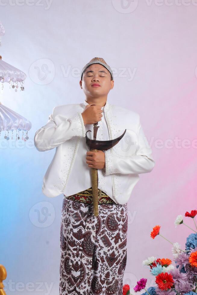 The gallantry of an elegant young Asian boy standing taking a keris up wearing modern Javanese traditional clothing, white velvet bridal kebaya on a white studio background. for advertising, fashion photo