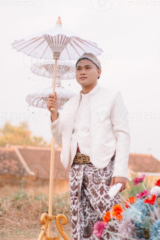handsome young Asian male standing candidly holding an umbrella wearing modern Javanese traditional clothing, white velvet bridal kebaya in an outdoor barren garden. for advertising, beauty, fashion photo