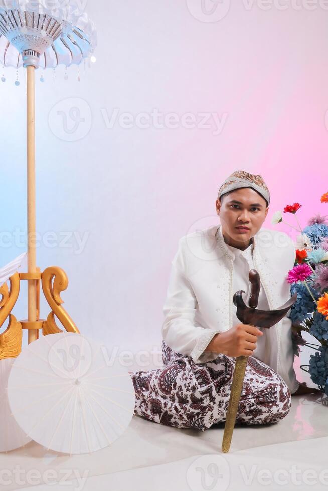 full body gallantry young elegant Asian man sitting holding a keris wearing modern Javanese traditional clothing, white velvet bridal kebaya on a white studio background. for beauty, fashion photo