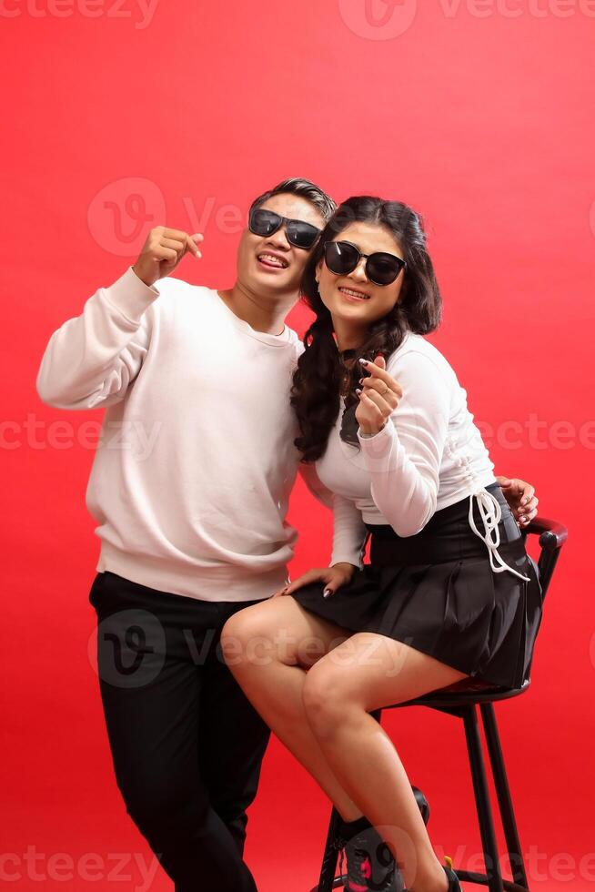 A pair of young lovers, a cheerful Asian man and woman wearing white sweaters, sunglasses, the woman sitting on a chair next to the man while showing a sign of saranghaeyo love. for romance, photo