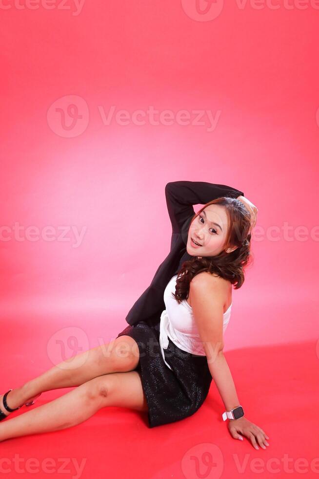 full portrait of a young indonesia woman in a career office sitting exclusively wearing a black suit, hands to the back of the head and the floor, for corporate, fashion, commercial, agency content photo