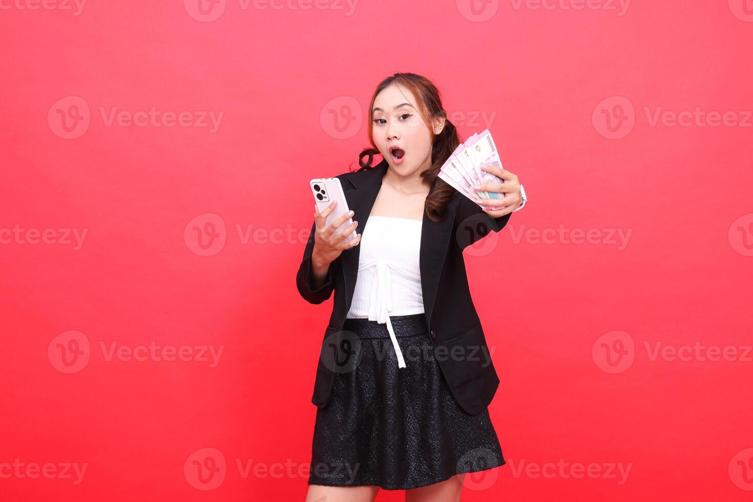 expression of surprised young indonesia career woman wearing formal black suit holding rupiah banknotes and cellphone in front, for technology, business, advertising, financial content photo