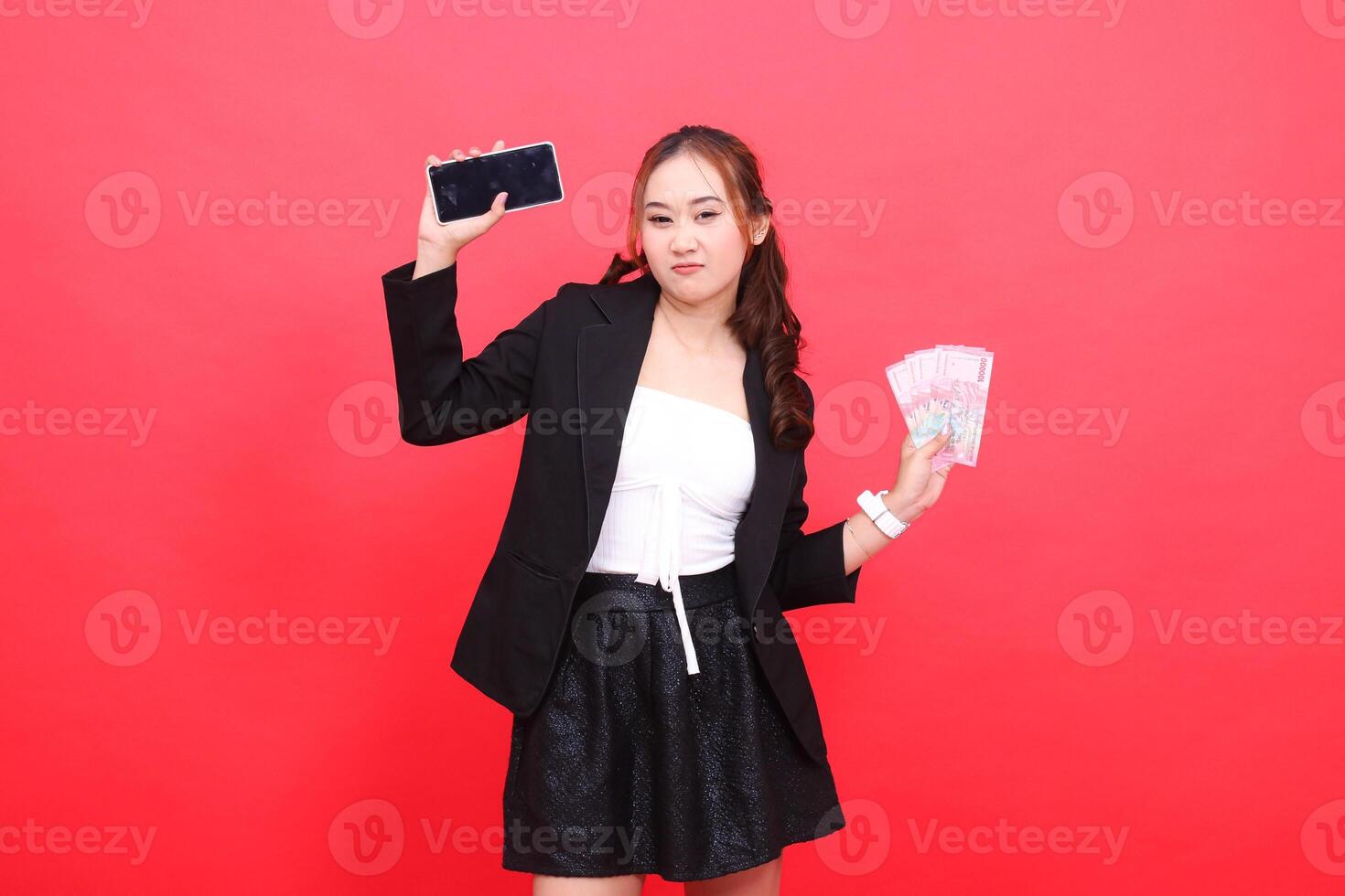 gesture of young Asian career girl frowning wearing black formal suit, hand holding rupiah money and cell phone up, for technology, business, advertising, financial content photo