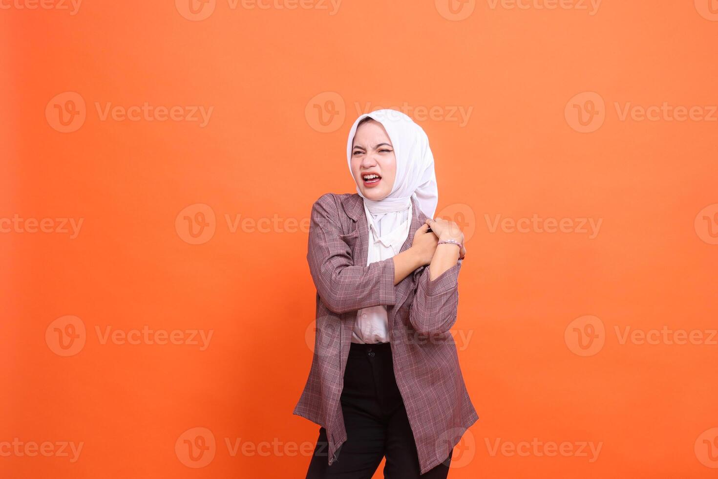 Portrait of a beautiful young Asian girl in pain grimacing in a hijab candid top, Experiencing back shoulder pain, hand position on shoulder with orange studio background. health care, commercial photo