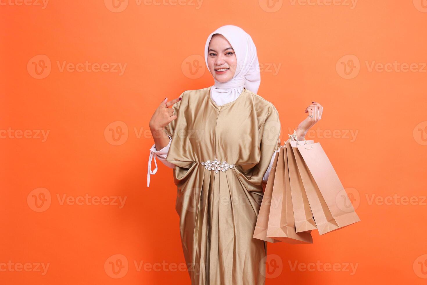 Cheerful young indonesia woman carrying a brown paper bag containing shopping items while pointing at it wearing a kaftan Muslim dress with a hijab. Concept of fashion, transaction, promotion photo