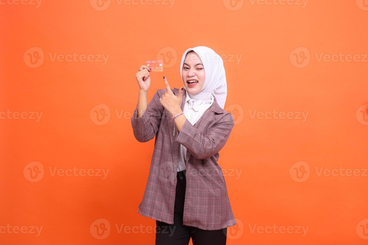 mature indonesia woman hijab suit cheerful winking holding plastic debit credit card while pointing at it isolated on orange background. satisfied with service for financial , mobile transactions, photo