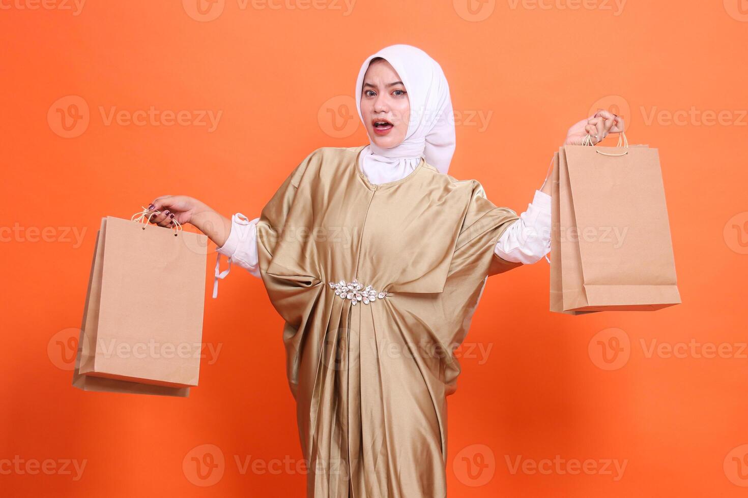 shocked young indonesia woman carrying aside a paper bag containing groceries wearing a kaftan Muslim dress with a hijab, isolated on an orange background. Fashion concept, transaction, promotion photo