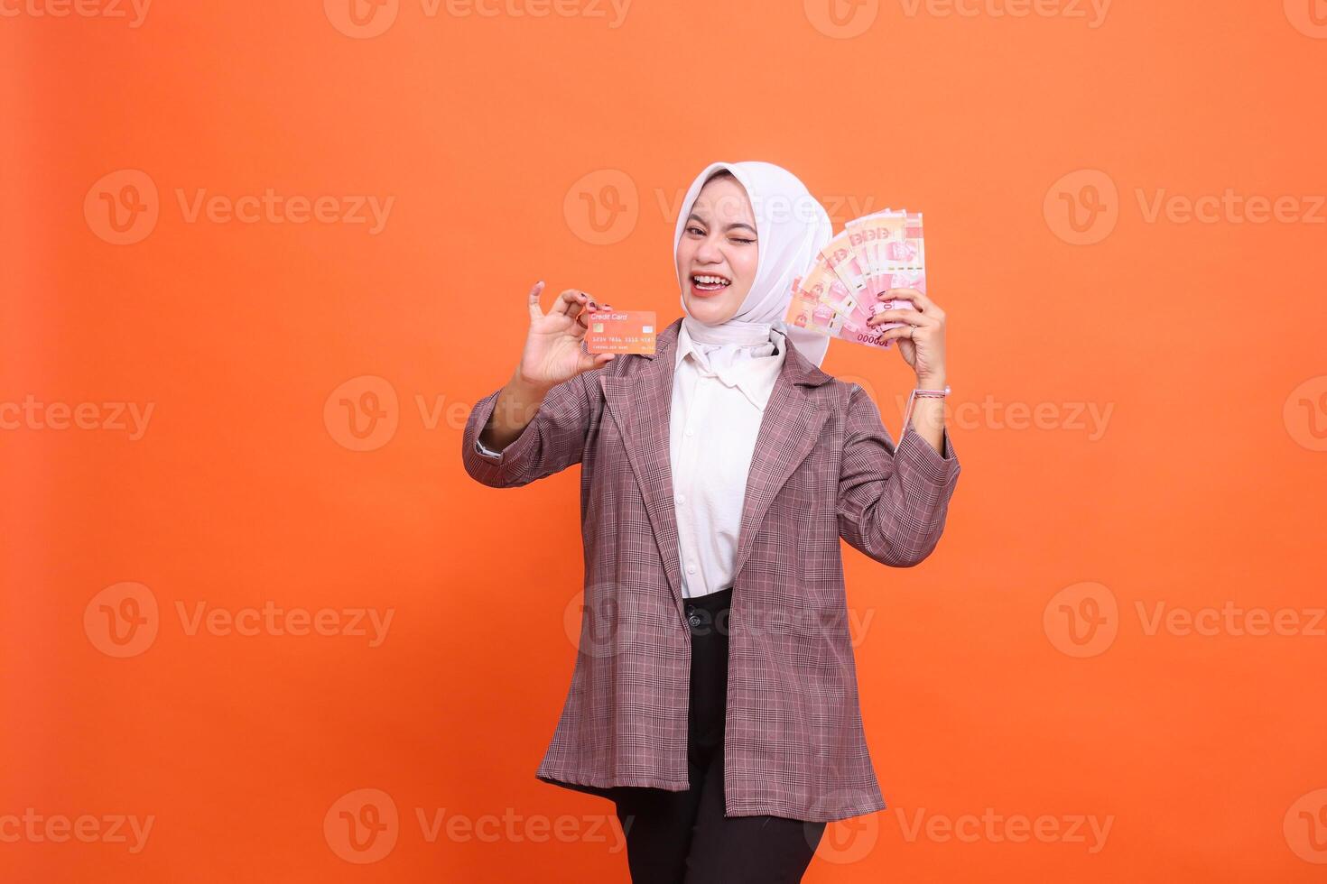 Young happy 20s year old indonesia girl winking wearing hijab office wear holding debit credit card and rupiah money in fan isolated on plain pastel orange background studio portrait photo