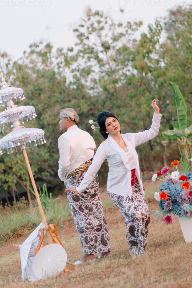 A pair of young indonesia male and female lovers standing, the woman dancing behind the man wearing modern Javanese traditional clothing, the bride's white velvet kebaya in an outdoor barren garden. photo