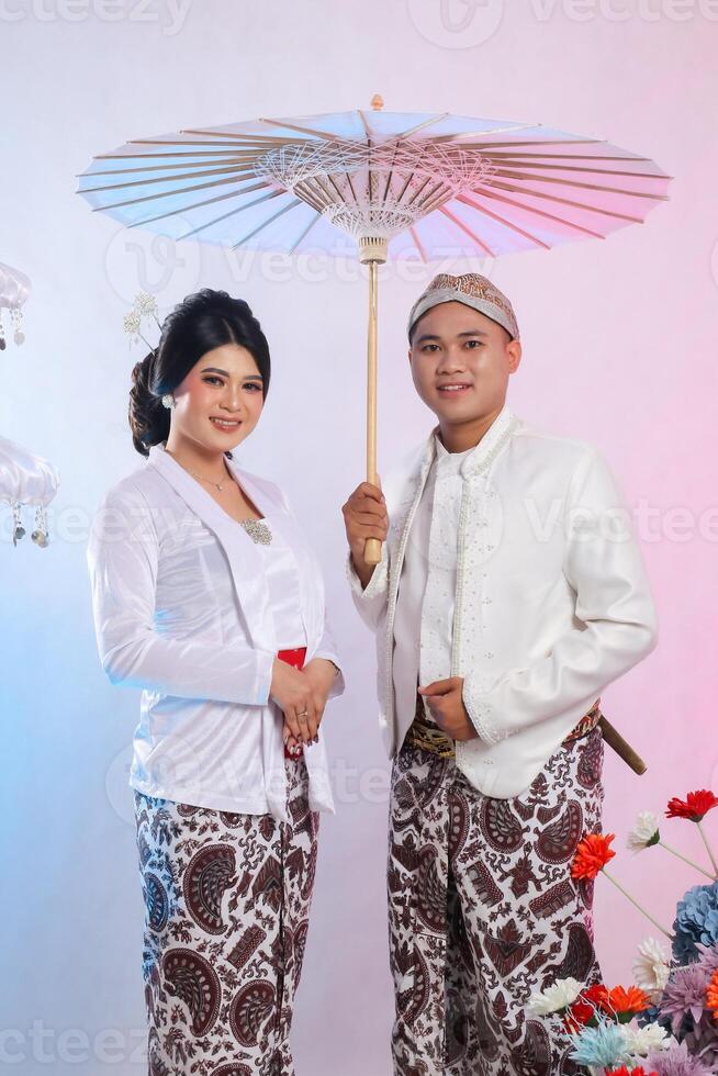 full body of a young Asian male and female lover standing smiling, man holding an umbrella wearing modern Javanese traditional clothing, white velvet for the bride and groom on a white background photo