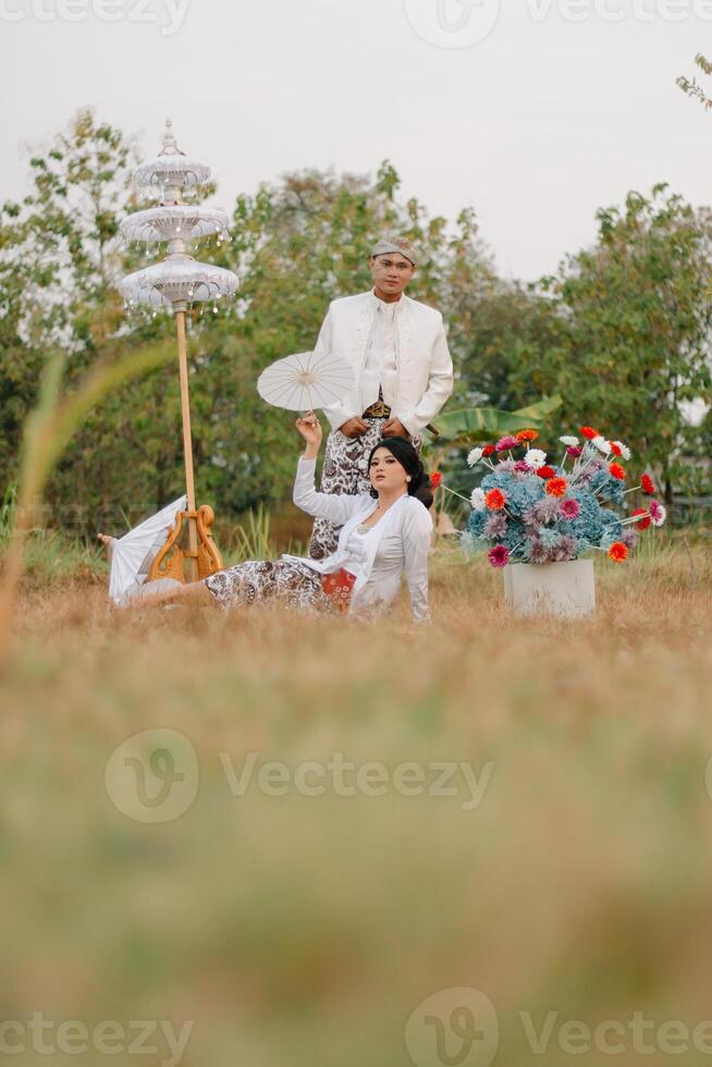 couple young indonesia male and female lovers smiling, women holding umbrellas sitting under modern Javanese traditional clothing, bridal white velvet kebaya in an outdoor barren garden photo