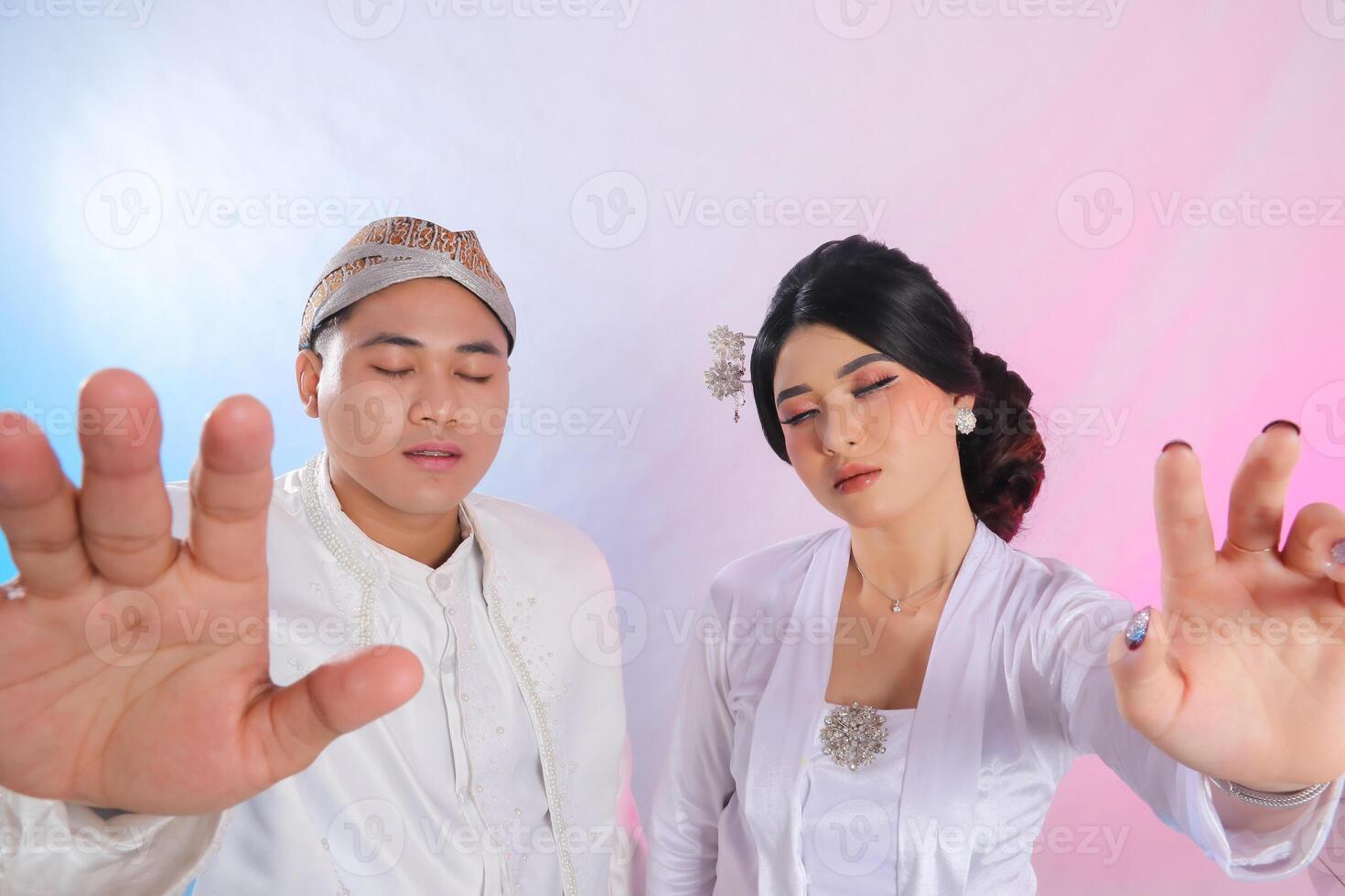 A pair of indonesia men and women with their eyes closed, hands facing forward, wearing modern Javanese traditional clothing, white velvet bridal kebaya, wedding concept with a white studio background photo
