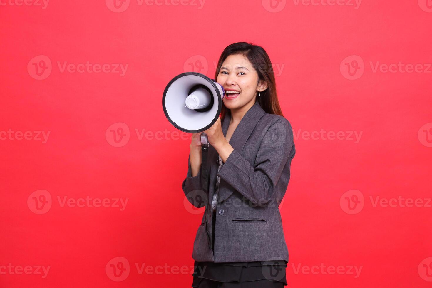 the expression of a cheerful Asian office woman shouting using a megaphone in front of her wearing a gray jacket and skirt on a red background. for broadcast, business and advertising concepts photo