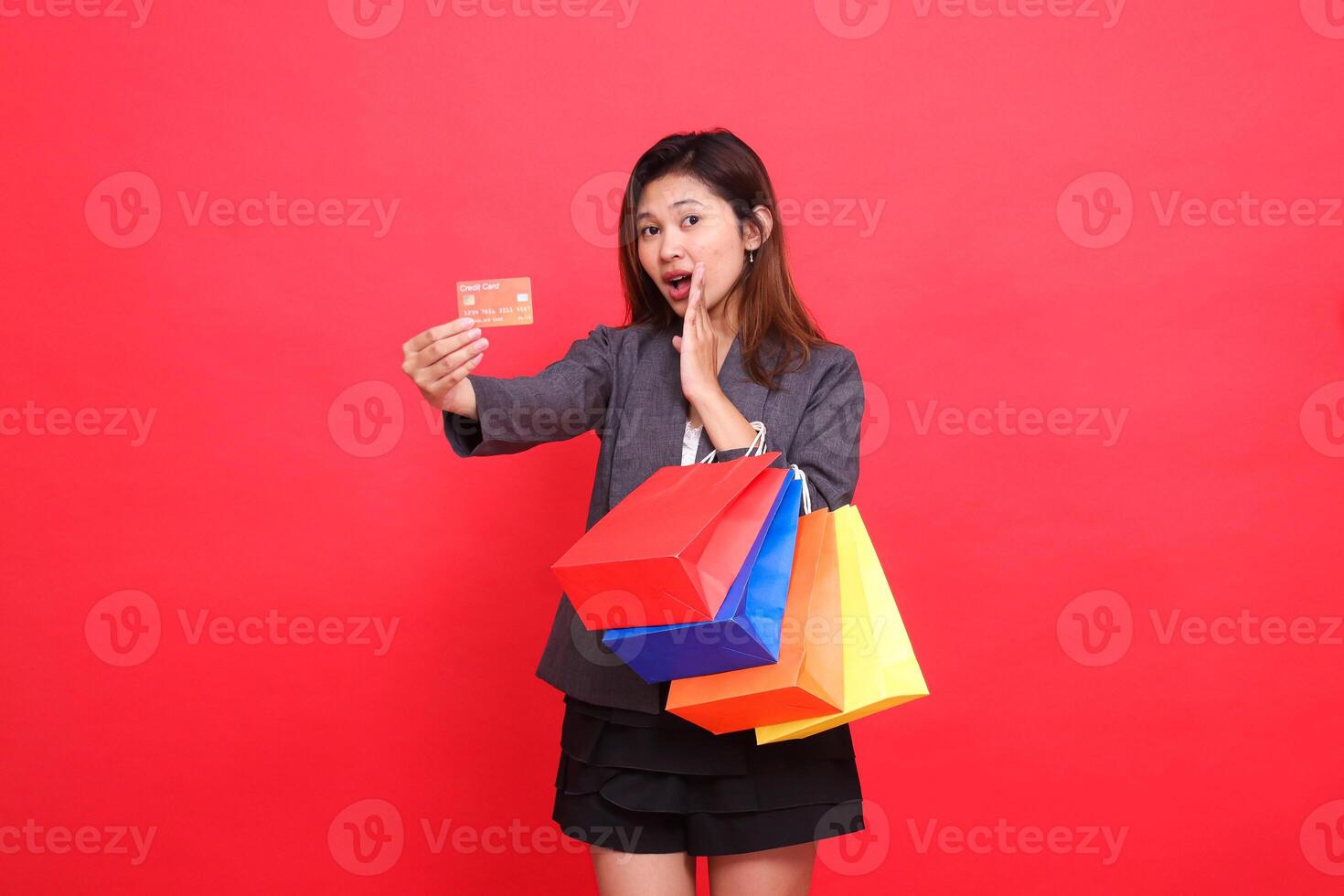 Asian office woman shocked to the left carrying a shopping paper bag while holding a credit debit card to the front wearing a formal jacket with a red background. for transaction and business photo