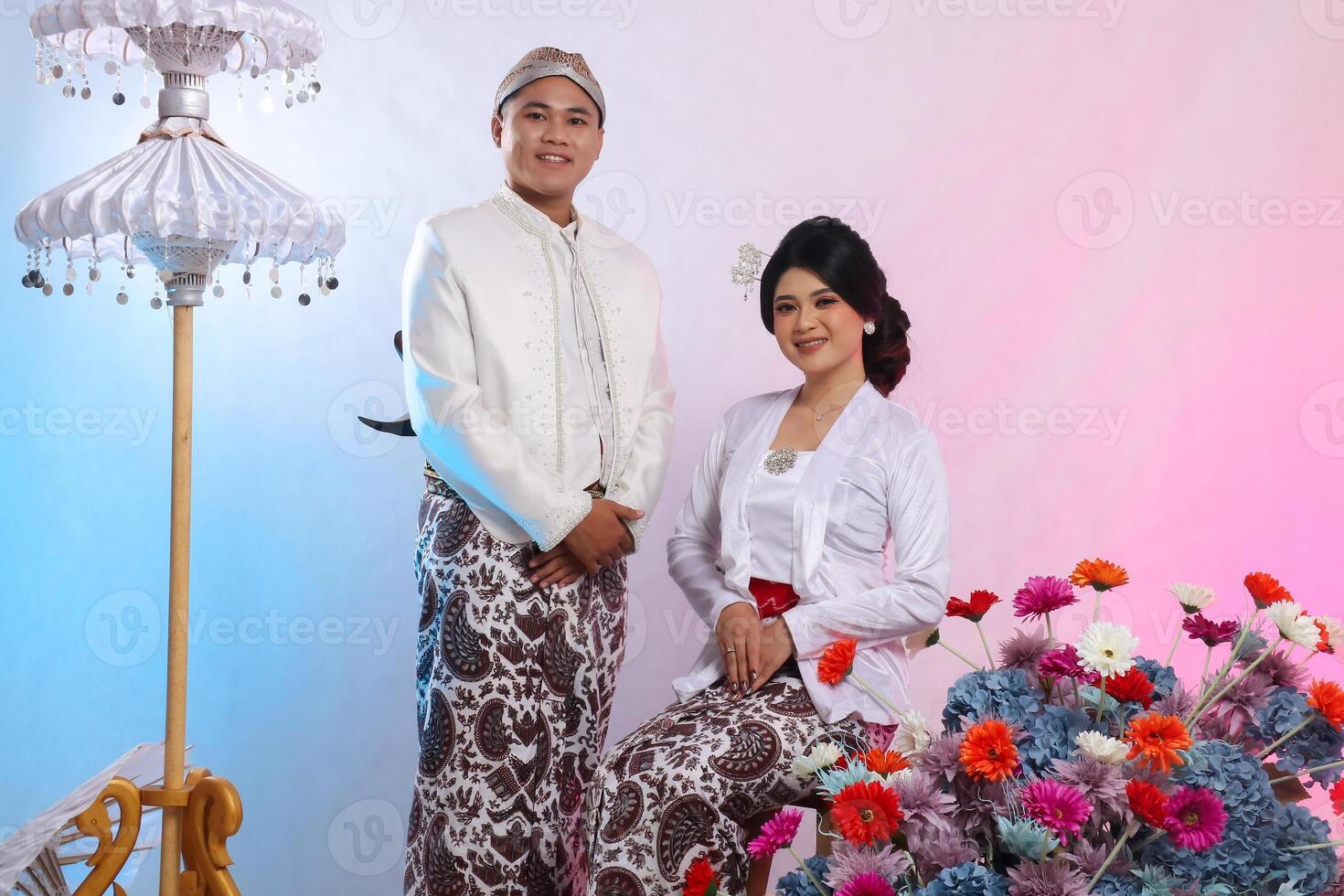 full body couple boy and girl asian woman smiling gracefully sitting next to man wearing modern Javanese traditional clothing, white velvet kebaya for bride and groom on a white studio background photo