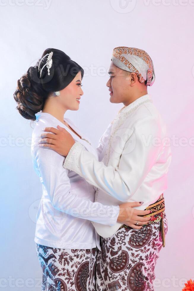 A pair of young indonesia men and woman lovers, standing smiling romantically facing each other wearing modern Javanese traditional clothing, white velvet bridal kebaya on a white studio background photo