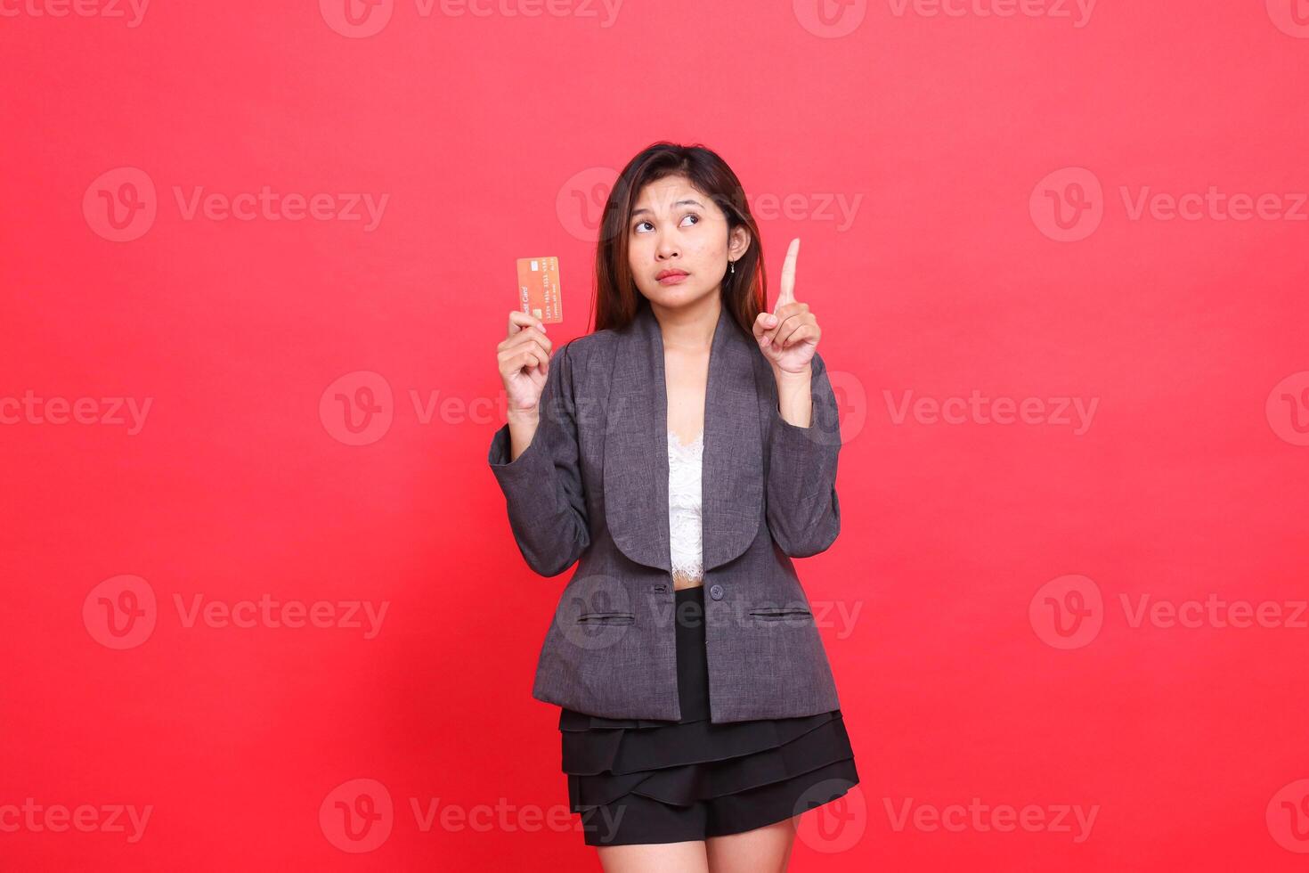 chinese office woman's expression shocked with her hand holding a debit credit card while pointing upwards, wearing a jacket and skirt on a red background. for financial, business and advertising photo