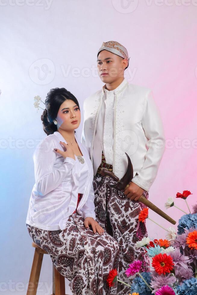 A pair of young indonesia men and women sitting elegantly sideways next to a man carrying a keris wearing modern Javanese traditional clothing, a white velvet bridal kebaya on white studio background photo