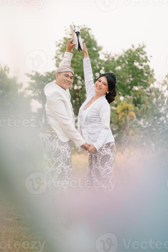 A pair of young indonesia male and female lovers walking, both arm in arm facing back happily wearing modern Javanese traditional clothing, white velvet bridal kebaya in an outdoor barren garden photo