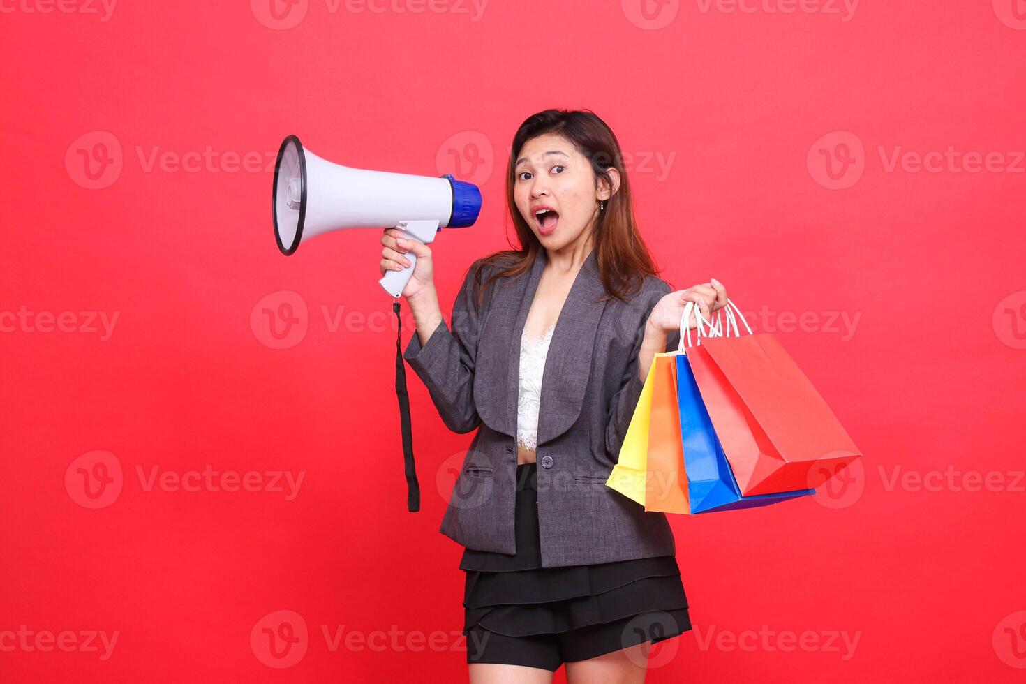 Asian sales woman shouts cheerfully at the camera through a megaphone speaker and holds up a shopping paper bag wearing a gray jacket on a red background. for transaction, lifestyle and business photo