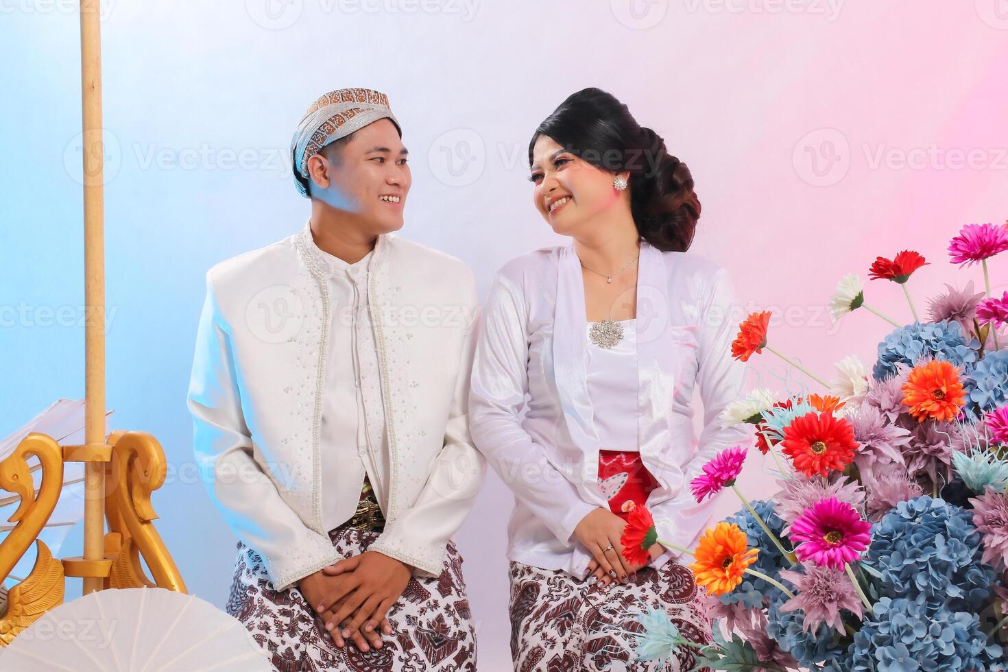 full body of a pair of cheerful indonesia boy and girl looking at each other sitting formally on the floor wearing modern Javanese traditional, white velvet bridal on white background photo