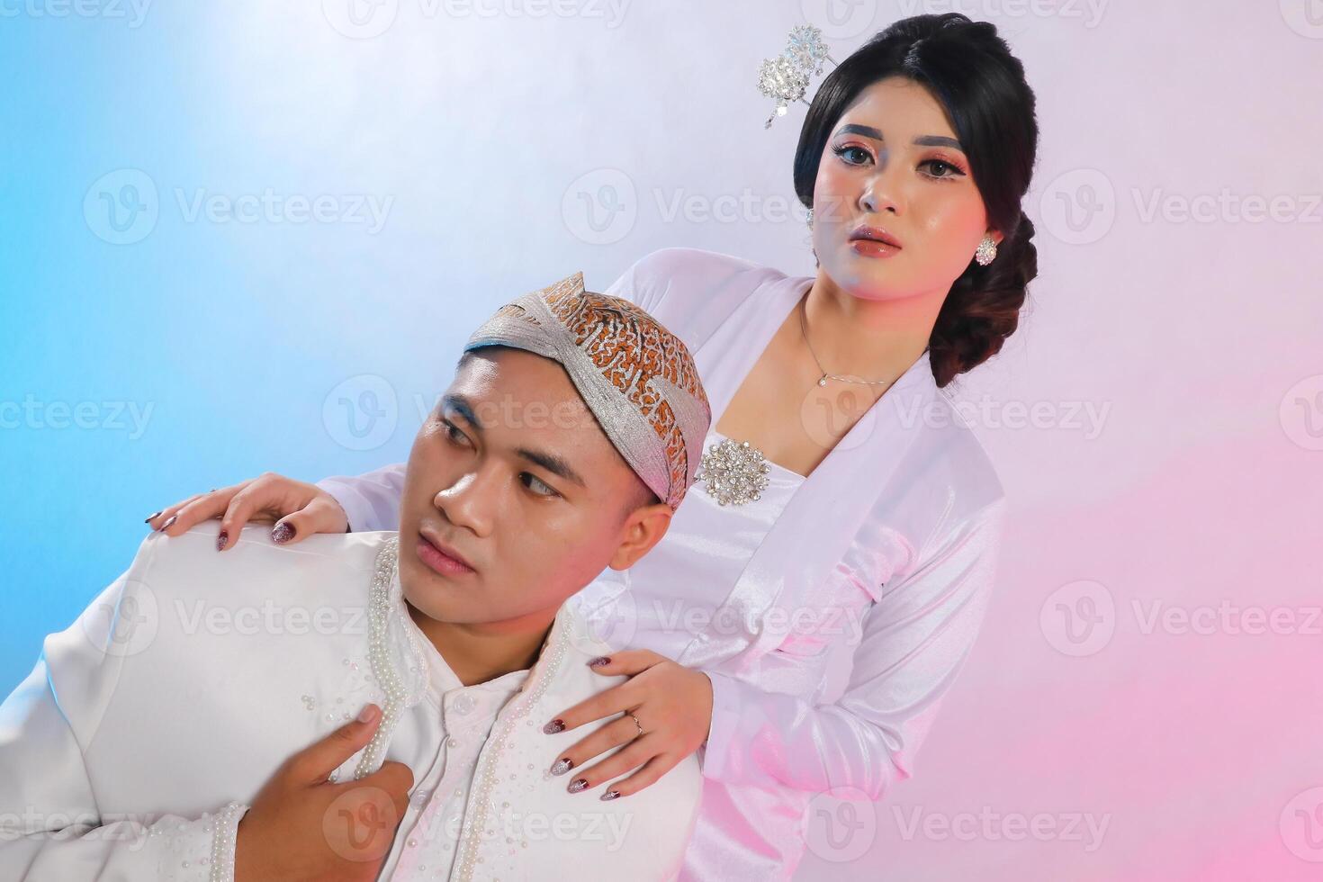 A pair of young Asian male and female lovers sitting candidly next to the woman who is wearing modern Javanese traditional clothing, a white velvet bridal kebaya on a white studio background photo