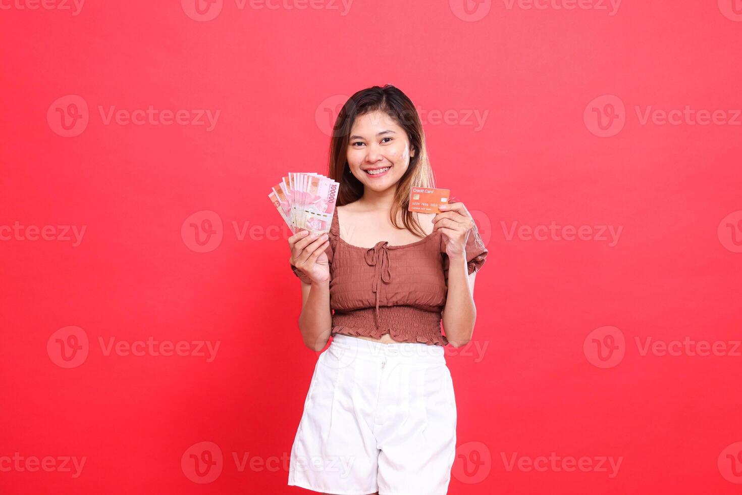 Asian woman smiling holding rupiah money and debit credit card wearing brown blouse top with red background. for transaction, technology and advertising concepts photo