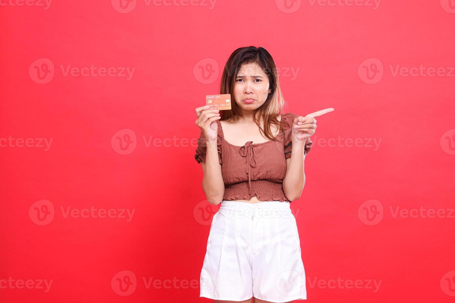 gesture of a sad, disappointed indonesia woman holding a debit credit card while pointing left, wearing a brown blouse on a red background. for transaction, technology and advertising concepts photo