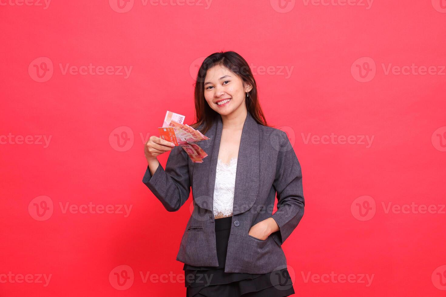 Cheerful Asian office woman expression holding debit credit card and money and putting money in pocket for camera wearing gray jacket and red skirt. for transaction, business and advertising concepts photo