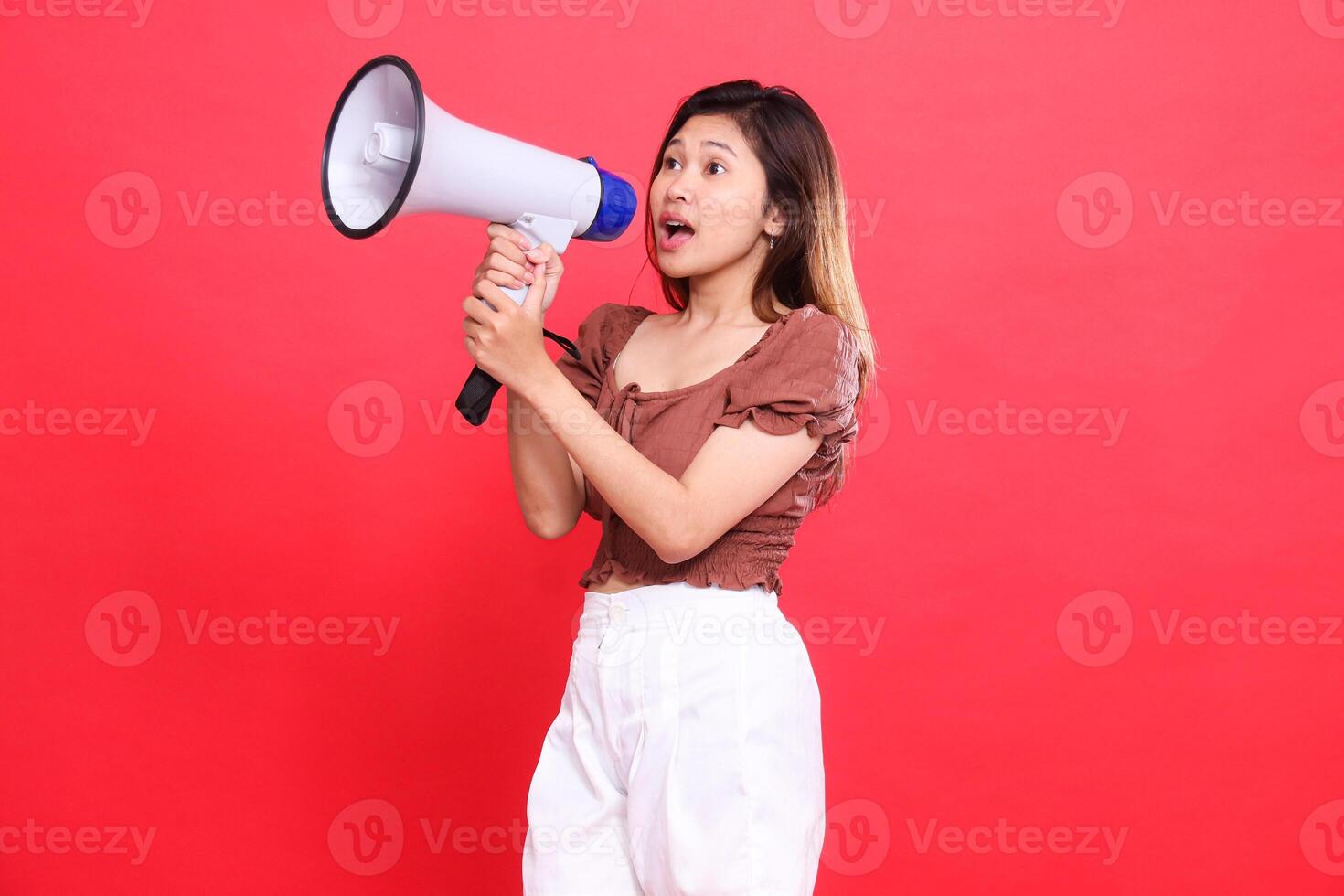 Elegant candid indonesia woman's expression shouting with both hands holding an audio megaphone wearing a brown blouse top and shorts on a red background. for broadcast, technology and advertising photo