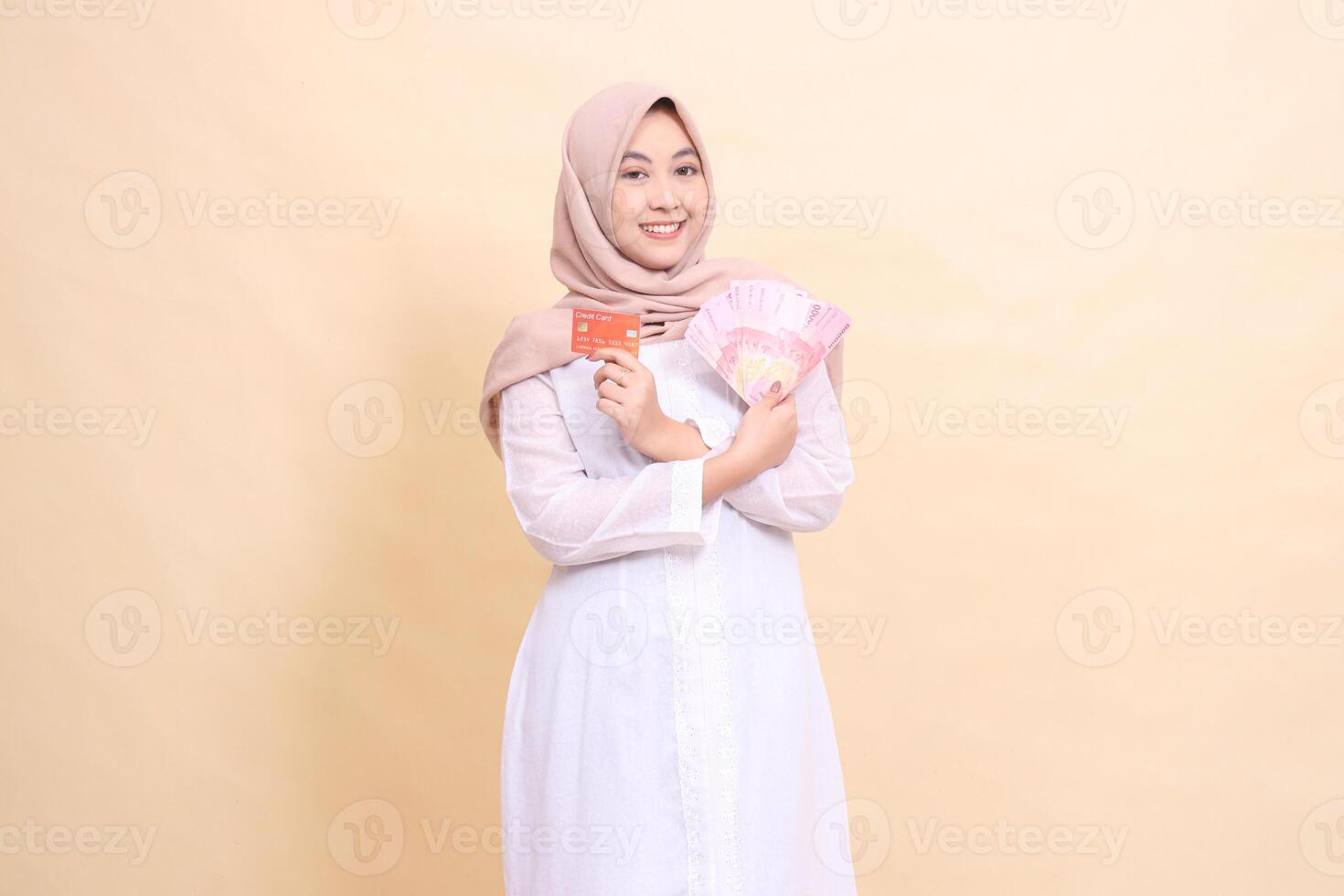 Asian Muslim woman in hijab smiling cheerfully holding rupiah cash and credit cards in crossed hands. Lifestyle, religious, business and finance concepts photo