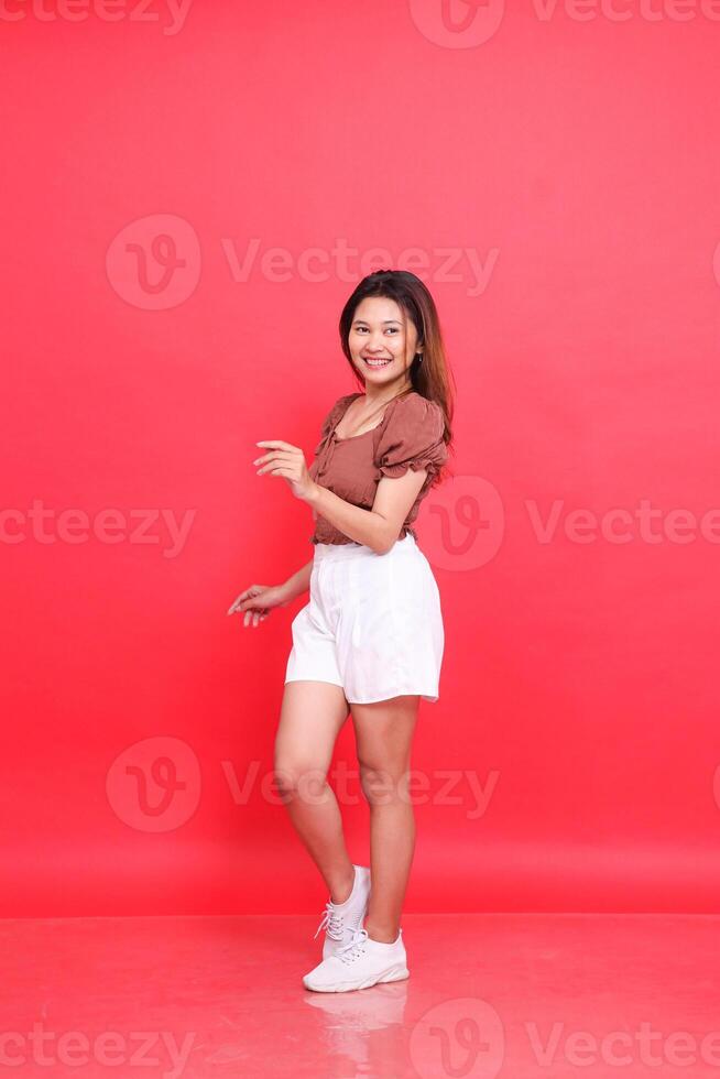 the charm of a cheerful indonesia woman in a candid dancing pose wearing a brown blouse and shorts on a red background. for fashion, lifestyle and clothing concepts photo
