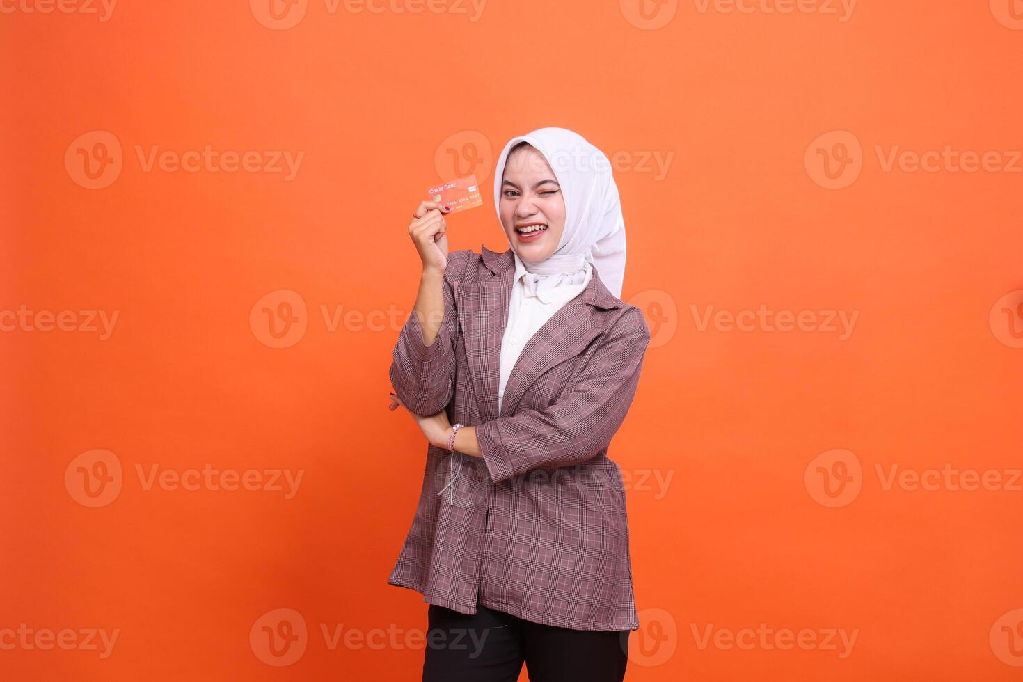 Young indonesia woman in hijab suit cheerful winks arms crossed while showing isolated plastic debit credit card on orange background. customer satisfied with service for financial ,mobile transaction photo