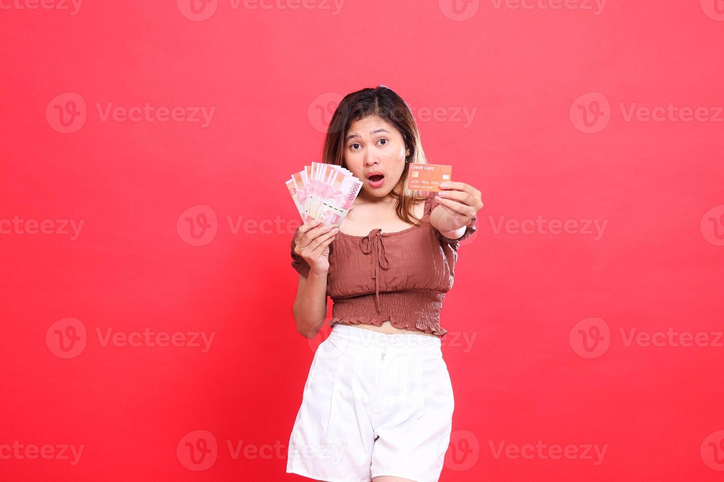 Shocked Asian woman holding Rupiah currency and debit credit card in front of her wearing a brown blouse with a red background. for transaction, technology and advertising concepts photo