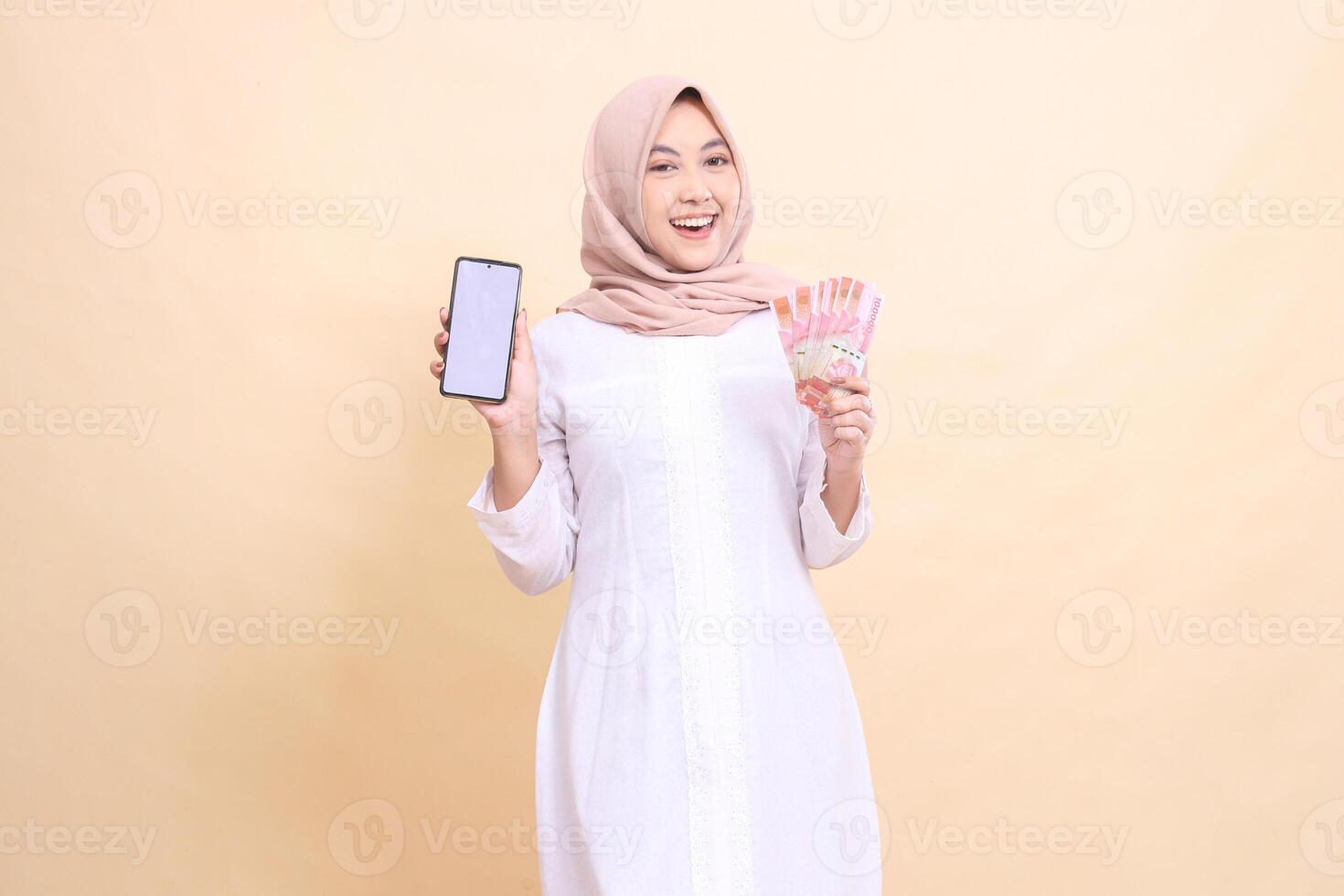 woman indonesia muslim young wearing a hijab smiles happily holding a cell phone gadget and rupiah money in her hand. Lifestyle, business and commercial concept photo