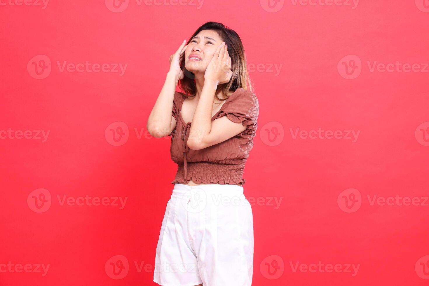 indonesia woman's expression with both hands holding her head up, very painful, dizzy, candid, wearing a brown blouse and shorts with a red background. for health, care and advertising concept photo