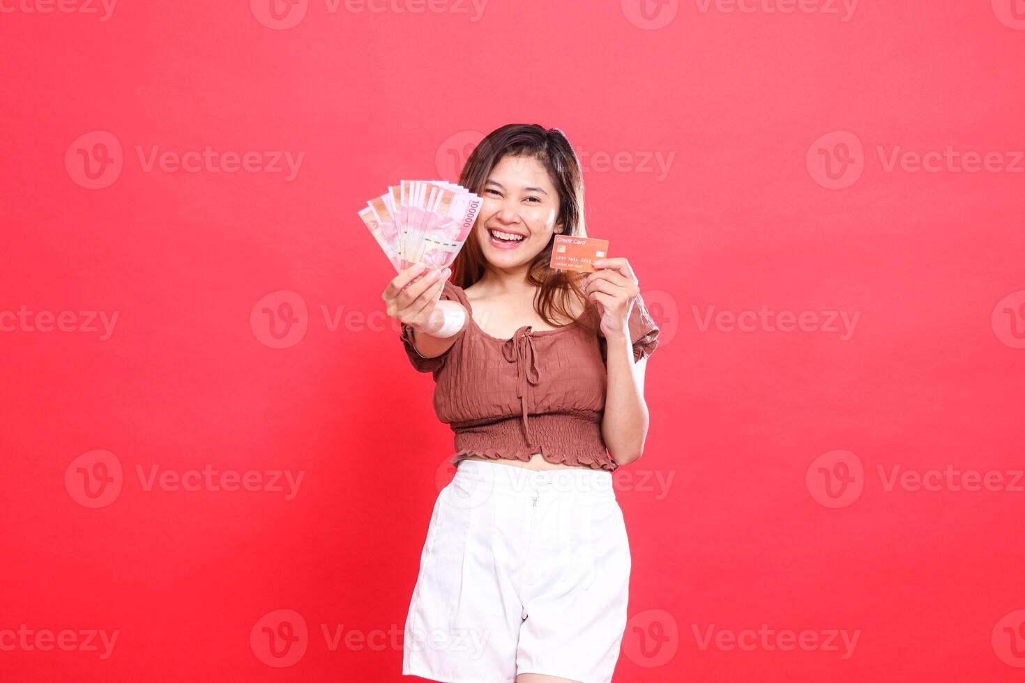 Happy indonesia woman holding rupiah money in front of her and a debit credit card wearing a brown blouse on a red background. for transaction, technology and advertising concepts photo