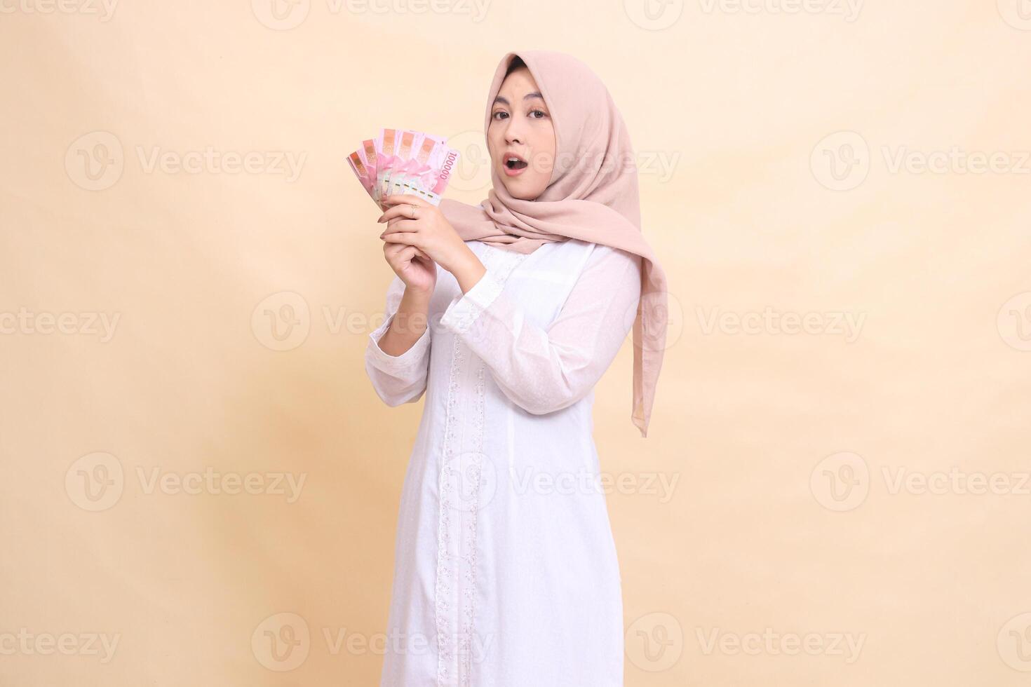 Muslim girl asian wearing a hijab slanted to the right is shocked holding rupiah cash in her hand. Lifestyle, religious, business and finance concepts photo