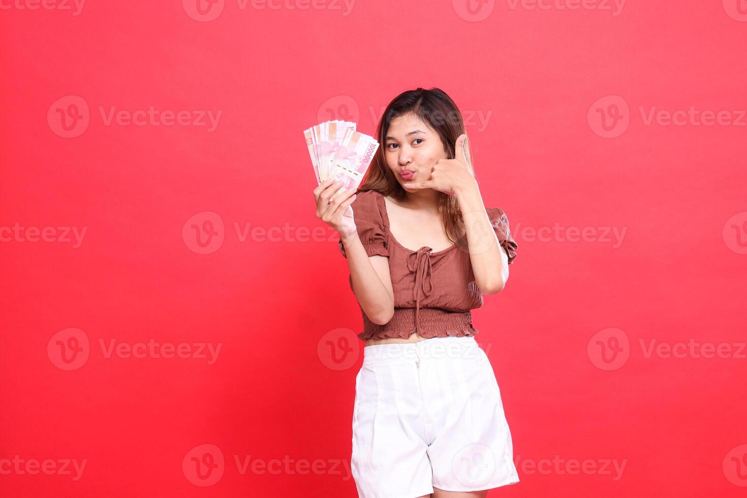 gesture of a cheerful young Asian girl holding rupiah money in front and a telephone sign. I am wearing a brown blouse with a red background. for transaction, technology and advertising concepts photo