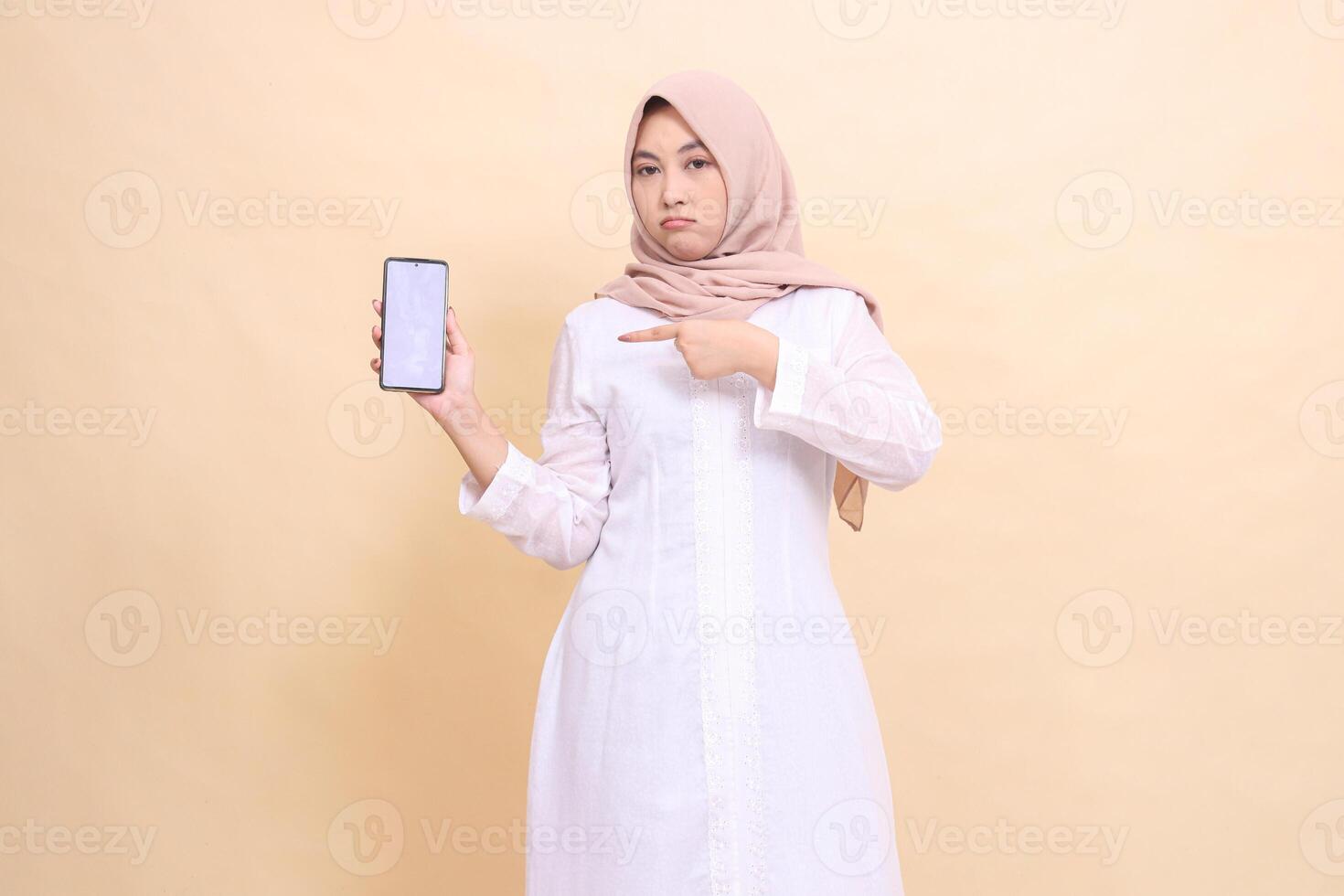 Young Asian Muslim woman wearing a hijab frowns holding her cell phone gadget to the right in her hand while pointing at it. Lifestyle, business and commercial concept photo