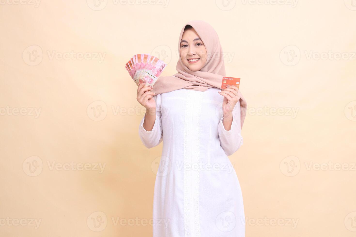 woman asian muslim wearing a hijab smiles cheerfully holding rupiah cash and credit cards in her hands. Lifestyle, religious, business and finance concepts photo