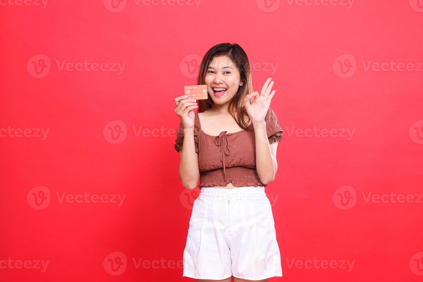 gesture of a cheerful indonesia woman holding a debit credit card with a zero sign okay, good wearing a brown blouse with a red background. for transaction, technology and advertising concepts photo