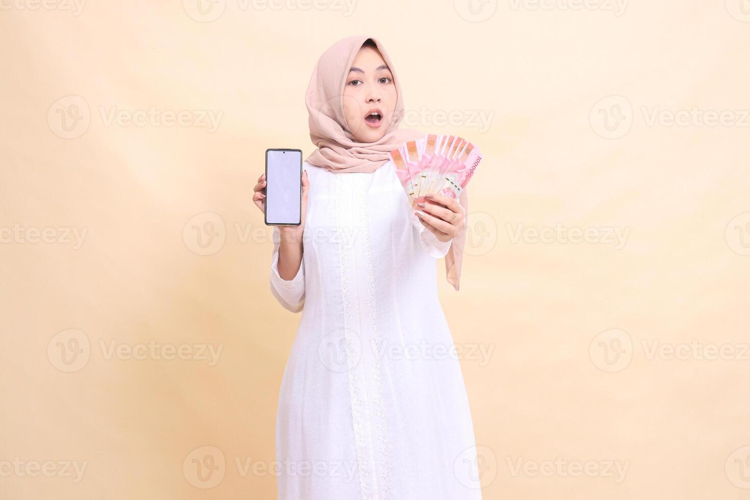 woman indonesia young muslim wearing a hijab is shocked holding a cellphone gadget and rupiah money in her hand. Lifestyle, business and commercial concept photo