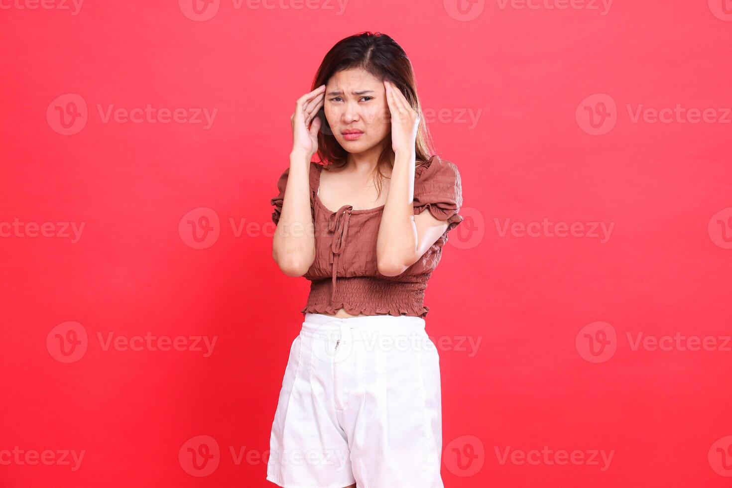 Asian woman's expression with both hands holding her head dizzy looking at the camera wearing a brown blouse top and shorts with a red background. for health, care and advertising concept photo