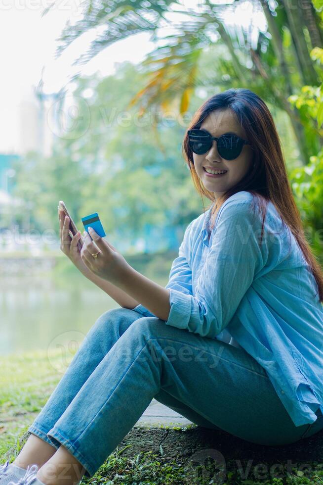 Beautiful Asian woman in sunglasses happy at the camera getting discount via credit card via cellphone gadget sitting on outdoor park grass. students for the themes of education and lifestyle photo