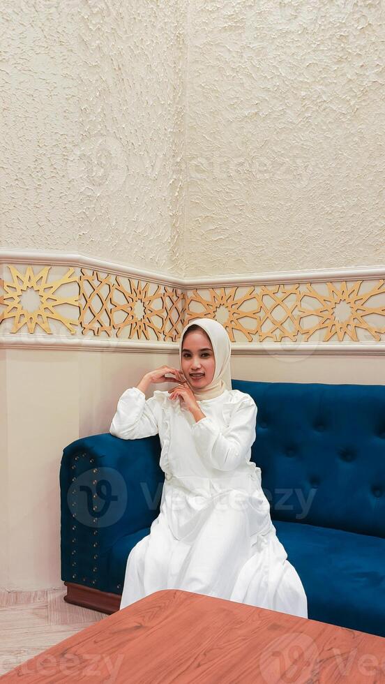 beautiful indonesia woman happy expression both hands face area and sitting on sofa with islamic ornament calligraphy background arabic atmosphere photo
